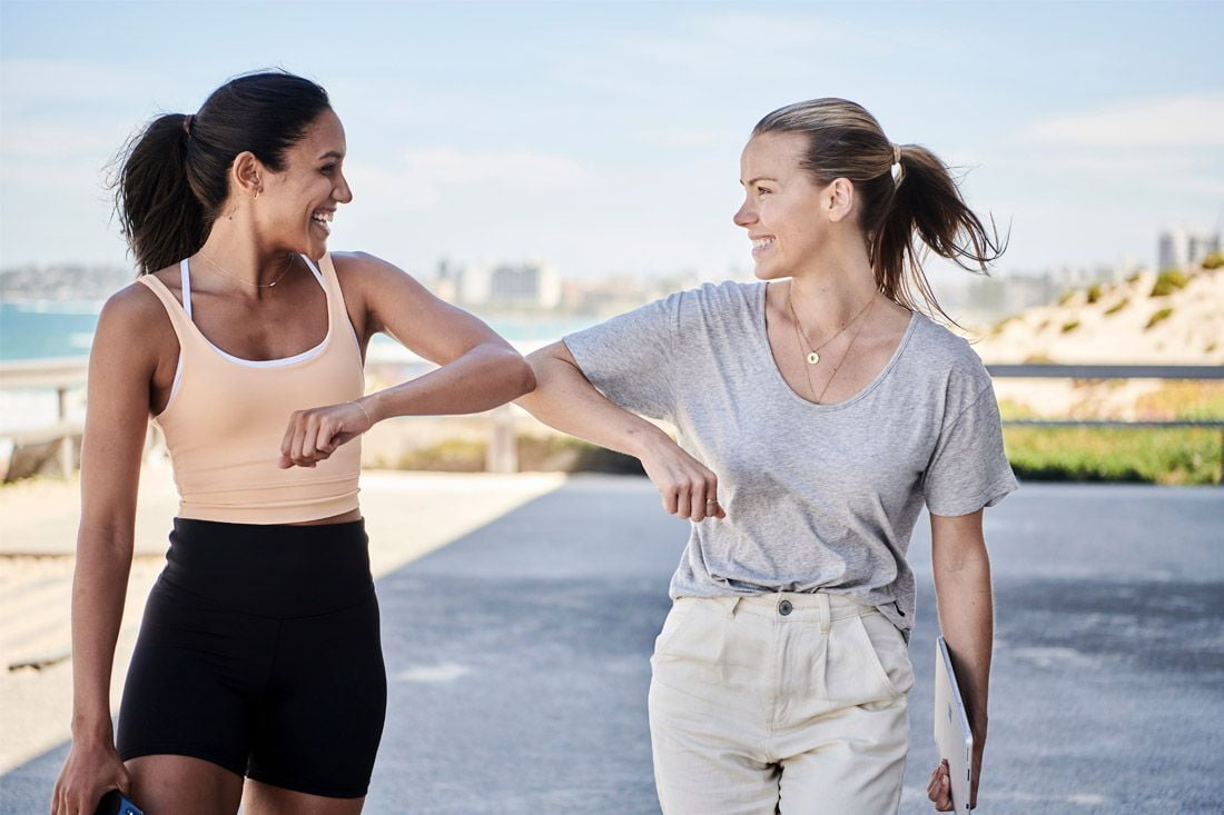 Two female friends are walking side by side in activewear, knocking elbows and smiling
