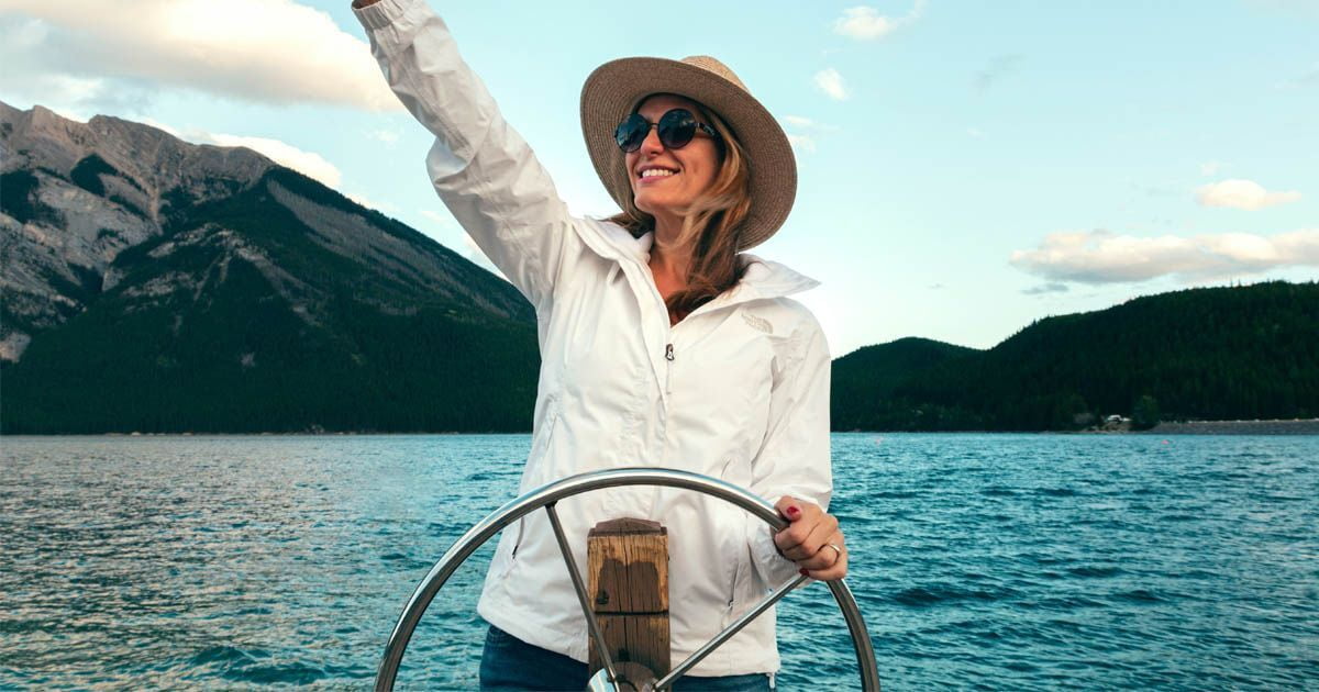 Woman stands on a boat holding onto the steering wheel, waving out into the distance