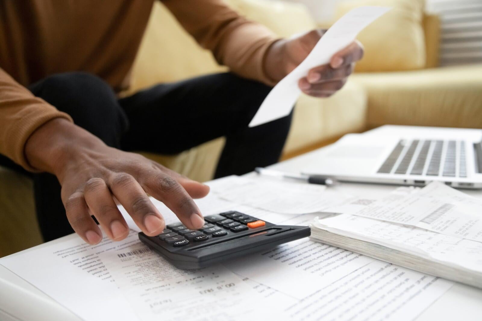 Man sits on a yellow couch bent over a coffee table covered in paper, holding a receipt in one hand and using a calculator in the other 